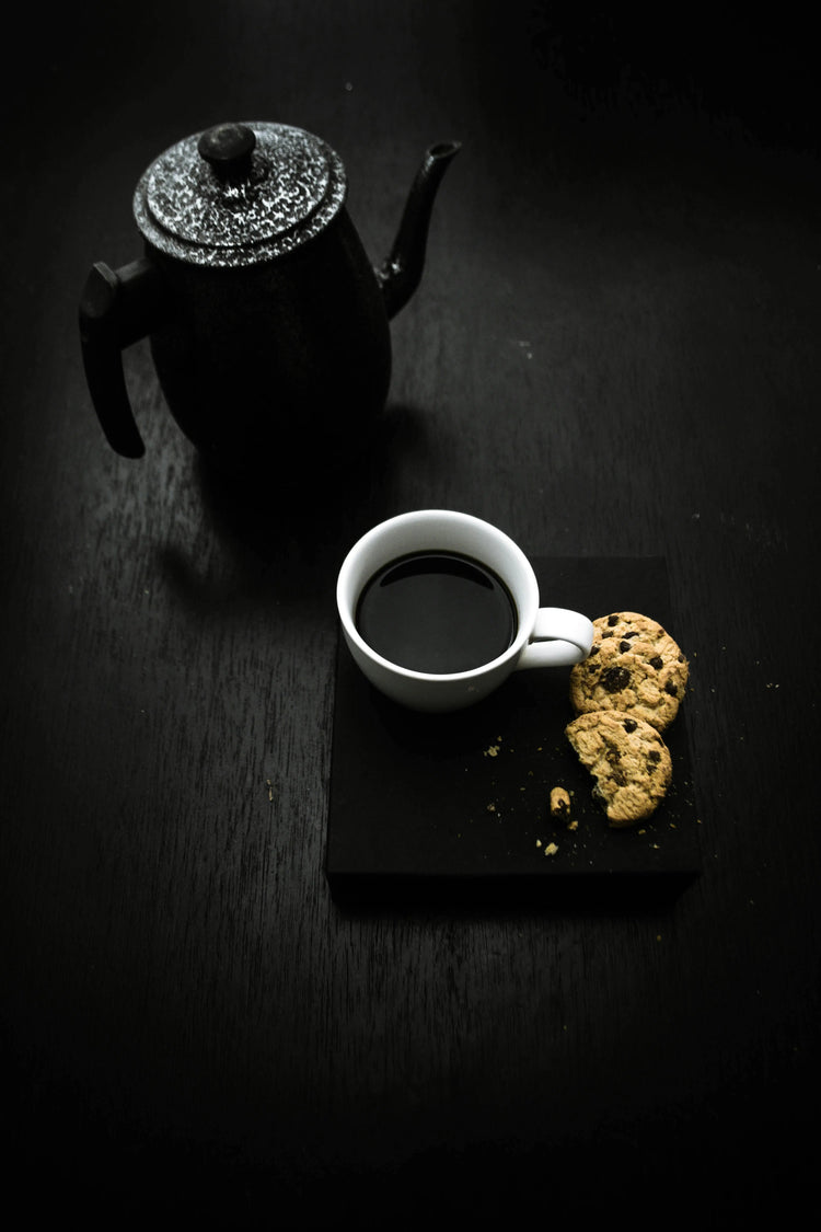 black wooden counter with black coffee ern next to white coffee mug and cookies